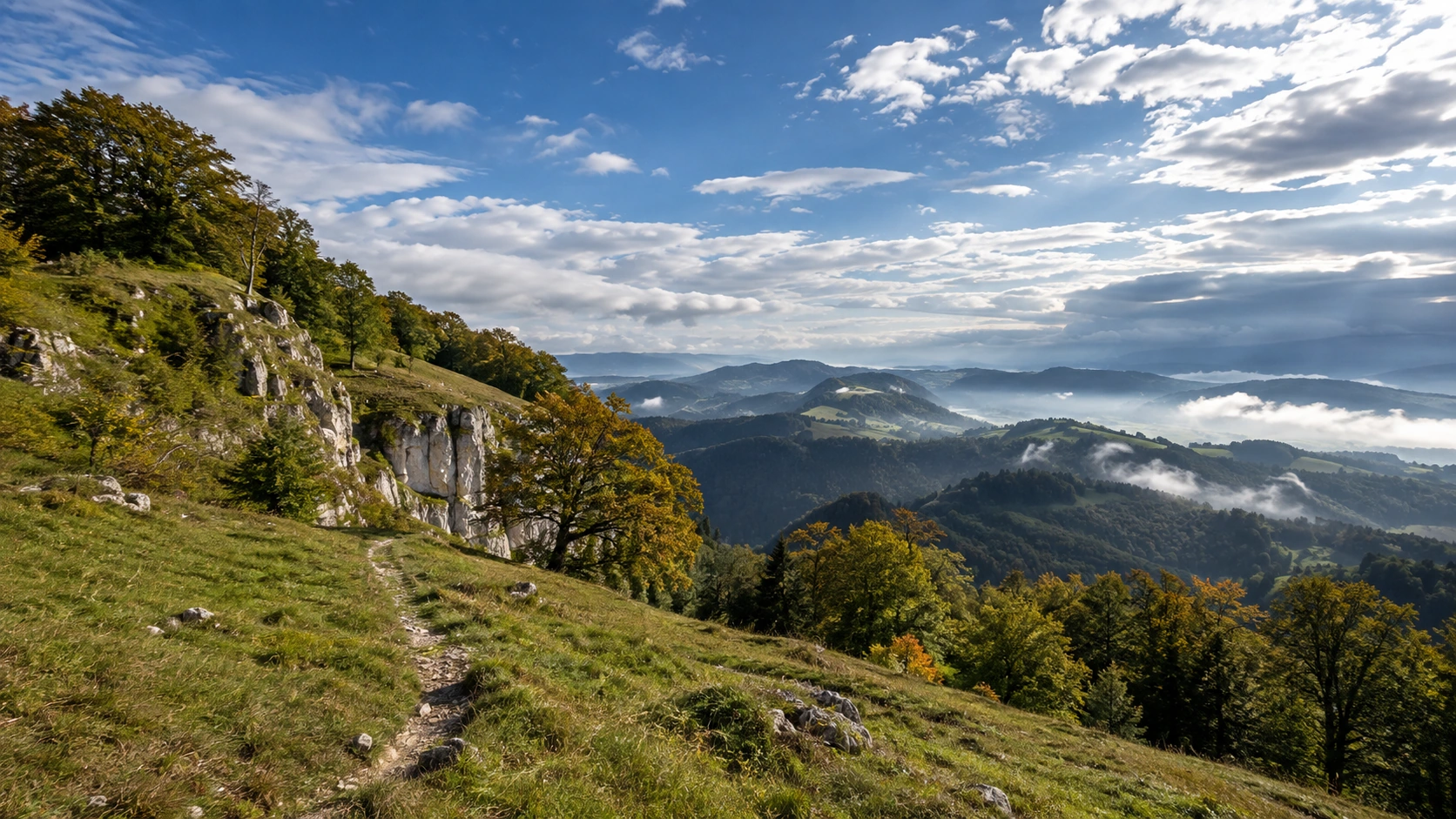 Grüne Jurahöhen mit Felsband, Wald, schmalem Bergweg und aufgelockerter Bewölkung.