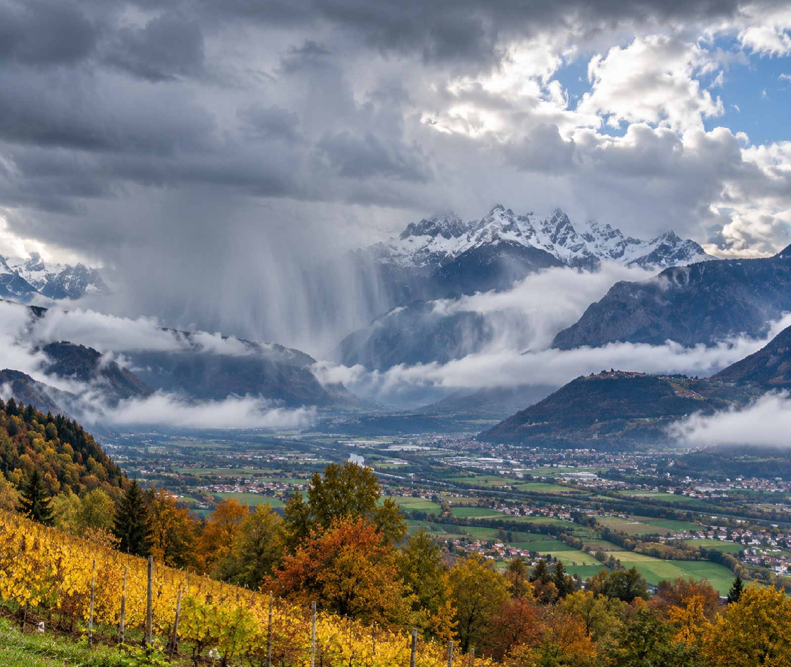 Herbstliche Alpenlandschaft mit Regenstaffeln, tiefen Wolken und frisch verschneiten Hochgipfeln als Symbolbild für den späten Temperaturrückgang.