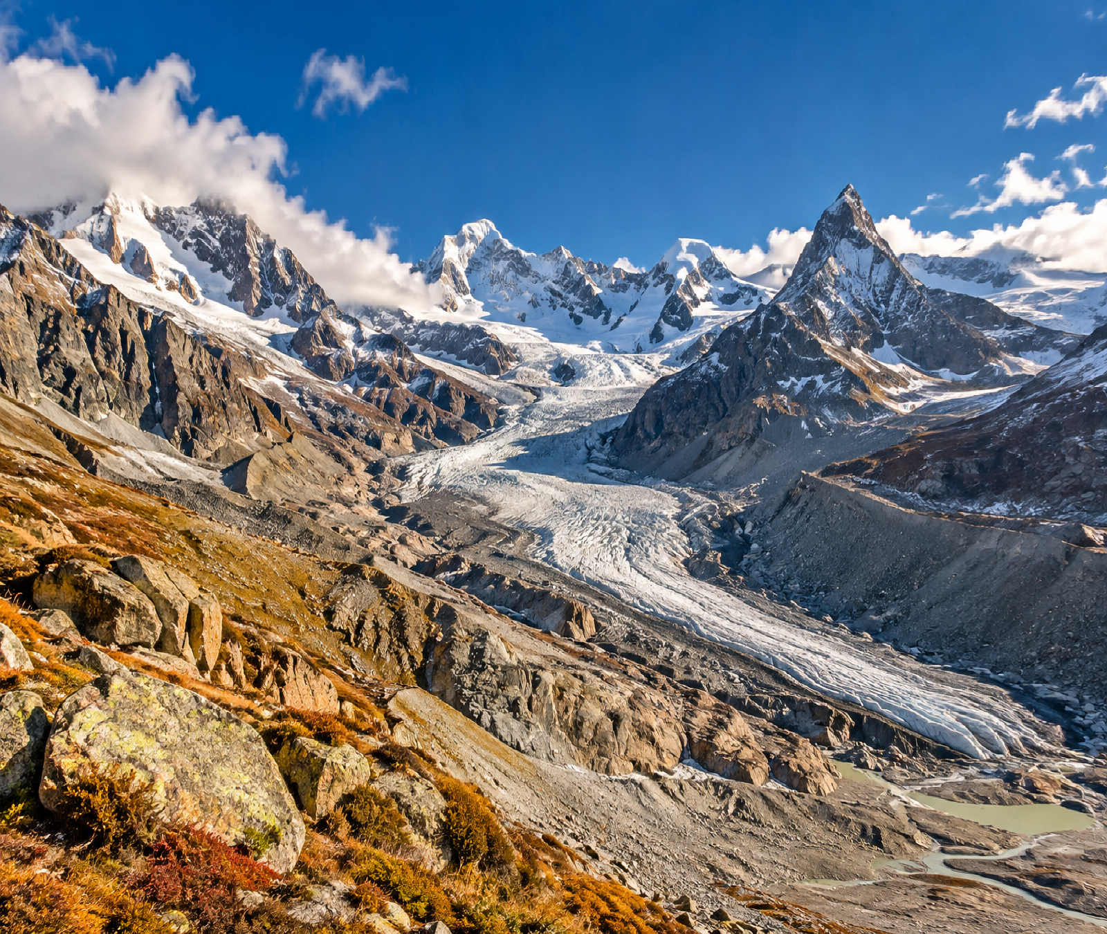 Symbolbild eines stark zurückgezogenen Alpengletschers mit Moränen und schneebedeckten Hochgipfeln im Herbst.